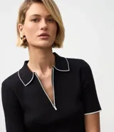 Blusa de punto negra de manga corta, ajustada al cuerpo, con cuello polo y ribetes blancos contrastantes. Presenta un cierre frontal con cremallera en el escote.