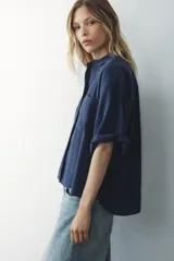 Camisa azul de cuello solapa y manga corta con dobladillo. Tiene dos bolsillos de parche en el pecho, bajo asimétrico con aberturas laterales y cierre frontal con botones ocultos.