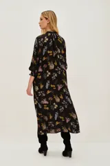 Vestido midi negro con estampado floral en tonos pastel y amarillos. Presenta escote pronunciado en V, mangas largas con puños anchos y detalles de bordado o pasamanería oscura en los bordes.