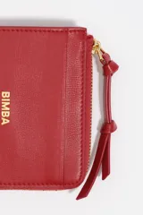 Monedero pequeño de estructura rígida, color negro, con cierre de cremallera y tirador de tejido amarillo mostaza. Presenta un bolsillo exterior frontal y el logo de la marca grabado en dorado.