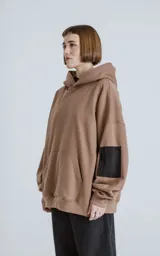 Buzo hoodie marrón de corte oversize, confeccionado en algodón rústico. Tiene capucha forrada, bolsillo tipo bomber con cierre en la manga y bolsillo canguro.