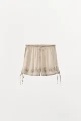 Shorts color crema de tiro alto con cintura elástica ajustable con lazada. Detalle de bordados florales combinados a tono. Aplicación de puntilla y abalorios. Bajo acabado con tira ajustable.