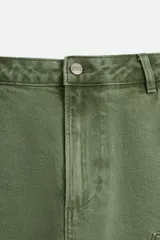 Bermuda color verde militar, de corte relaxed fit, con bolsillos frontales y traseros tipo plaqué, detalle de tira y bolsillo multifunción en la pierna, y bajo irregular.