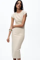 Vestido midi color beige confeccionado en hilatura con algodón. Tiene cuello redondo, manga corta caída y detalle de drapeado lateral.