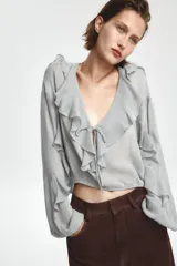 Blusa color marrón con escote en pico, manga larga abullonada con elástico en los puños y volantes en el escote y mangas.