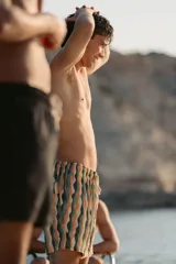 Short de baño para hombre con elástico en la cintura, tres bolsillos (dos laterales y uno trasero), tela con 10% spandex y secado rápido con doble costura de seguridad, interior con mesh hipoalergénico y accesorios waterproof. Diseño con rayas onduladas en tonos beige, verde, naranja y negro.