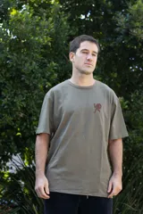 Remera color verde oliva con efecto gastado, de manga corta y cuello redondo. Presenta un pequeño estampado en el pecho de una figura humana estilizada en color marrón.