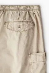Pantalón cargo de algodón color beige, de corte holgado y tiro medio. Presenta cintura elástica con cordón ajustable, bolsillos laterales y grandes bolsillos cargo con solapa en las piernas. Los bajos son ajustables con cordón oculto.