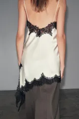 Vestido corto de satén color blanco, con tirantes finos, escote en pico y bajo asimétrico con abertura. Detalle de encaje negro en escote y ruedo.