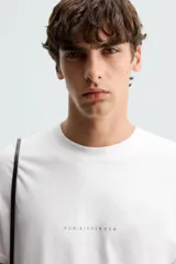Camiseta blanca de corte holgado, cuello redondo y manga corta. Estampado frontal con texto en color negro y parche en la espalda. Detalle de pliegues en la espalda.