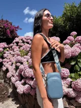 Bandolera pequeña tipo 'phone bag' de símil cuero color rosado pálido, con cierre de cremallera y correa ancha de tela con diseño de chevron en tonos claros.