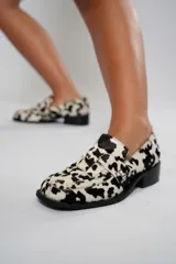 Mocasín de cuero con estampado animal print de vaca en tonos marrón y blanco, con capellada enrejillada y suela plana con plataforma delantera y taco bajo.