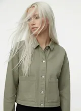 Chaqueta corta de algodón color verde oliva, con cuello camisero, cierre frontal de botones metálicos y dos bolsillos de parche en el frente. Presenta un corte relajado.
