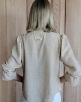 Blusa color beige con cuello con volados, mangas largas abullonadas con puños elásticos y cierre con botones en la parte delantera.