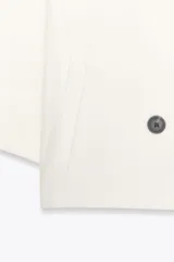 Blazer blanca de cuello solapa y manga corta con vuelta. Bolsillos delanteros de vivo y cierre frontal cruzado con botones.