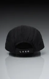 Gorra negra de estilo 5 paneles, con tejido acolchado o con costuras horizontales y visera plana. Presenta un pequeño parche cuadrado negro con un logo blanco bordado en forma de 'X' o lazo.