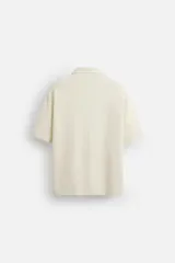 Camisa de manga corta color crudo, con cuello bowling, corte relaxed fit y cierre frontal con botones. Presenta apliques de parches con texturas combinadas a contraste en el delantero y aberturas laterales en el bajo.