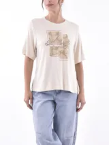 Remera blanca de manga corta con cuello redondo y estampado frontal que dice "SAVAGE" con una figura de animal (posiblemente un leopardo o jaguar) hecho de lentejuelas o material brillante. Debajo del estampado principal se lee "LIFE GOES ON ENJOY".