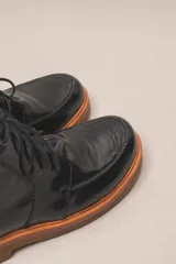 Bota de cuero vacuno con acabado charol, color marrón oscuro. Presenta un diseño de caña corta con cordones frontales y costuras decorativas en la puntera. La suela es de goma tipo crepe.