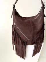 Bolso tipo hobo color tostado, hecho de cuero con flecos largos en el frente y dos bolsillos con cierre. Tiene una manija regulable con hebillas metálicas.