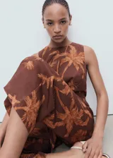 Mono largo sin mangas de color marrón con estampado de palmeras. Presenta un diseño con cuello redondo, detalle de nudo en la parte delantera y aberturas laterales tipo cut-out. Está confeccionado en una mezcla de lyocell y lino con forro de algodón, cuenta con perneras de corte recto y cierre de cremallera en la parte posterior.