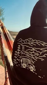 Buzo hoodie negro con capucha y cordones ajustables. Estampado en la espalda con diseño de paisaje de montaña, árboles, río y camioneta con canoa en color blanco.