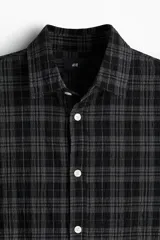 Camisa de popelina de algodón texturizado a cuadros negros y grises, de corte holgado, con cuello inglés, tapeta lisa, canesú con pliegue de caja en la espalda, mangas largas con abertura y cierre ajustable de botón en los puños, y bajo levemente redondeado.