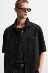 Sobrecamisa color beige de tejido elástico, con cuello solapa, manga corta y bolsillos de parche en el pecho. Cierre frontal con botones a presión.