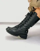 Borcego de cuero vacuno color negro, con suela de goma artesanal, caña alta con aplique capitoneado desmontable y horma de punta redonda.