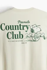 Remera color crema de corte holgado, con cuello redondo y estampado pequeño de Snoopy y la frase "Peanuts" en el pecho.