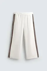 Pantalón jogger blanco con textura acanalada y corte amplio. Presenta cintura elástica y bandas laterales marrones. Incluye bolsillos laterales y trasero de parche.