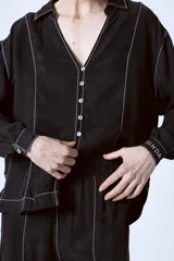 Blusa negra con botones, cuello camisero y mangas largas con puños. Presenta pespuntes blancos en contraste.