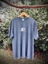 Remera oversize de algodón con efecto lavado (acid wash) en tono azul grisáceo. Presenta un estampado circular en la espalda con una palmera y texto que dice "SUNSET", "TROPICAL" y "PALM BEACH".
