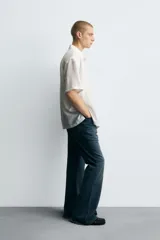 Camisa blanca de manga corta con cuello solapa y textura jacquard irregular. Corte relajado (relaxed fit) con aberturas laterales y cierre frontal de botonadura.