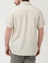 Camisa de manga corta color beige, con cuello clásico, cierre frontal con botones y bolsillo en el pecho izquierdo.
