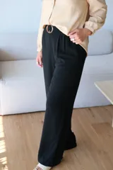 Pantalón negro de tiro alto con pliegues frontales y pierna ancha, ajustado a la cintura con un cinturón marrón claro con hebilla circular.