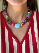 Collar corto con cuentas grandes y coloridas de formas variadas (redondas, ovaladas, planas) en tonos vibrantes como celeste, fucsia, amarillo, azul, rojo y marrón. Algunas cuentas tienen acabado liso y otras texturizado o jaspeado.