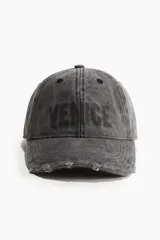 Gorra de lona de algodón color gris oscuro con estampado frontal de la palabra "VENCE" en letras grandes. Tiene detalles desgastados en la visera y ojales bordados. Cuenta con cinta antitranspirante de algodón y hebilla ajustable de metal en la parte trasera.