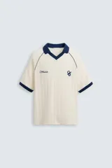 Polo color crema con cuello solapa azul marino, mangas cortas con ribete azul marino, bordado de texto en el pecho y bordado de logo en el abdomen.