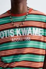Camiseta de algodón a rayas horizontales en tonos verde agua, naranja, marrón y blanco. Tiene cuello redondo y mangas cortas. Estampado frontal con el texto "OTIS KWAME" y "ACCRA 1986" en color blanco.