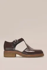 Zapato tipo guillermina de cuero charol color marrón oscuro, con tira T y hebilla ajustable en el lateral. Posee puntera cuadrada y suela de goma color caramelo con taco bajo.