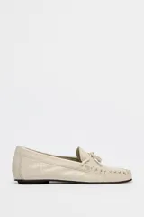 Zapato plano tipo mocasín color crema, confeccionado en cuero, con detalle de lazo con pasador y fruncido en la parte delantera. Pespunte en el corte y acabado en punta redonda.