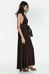 Vestido largo color marrón oscuro, con cuello alto y corte al cuerpo en la parte superior, y falda acampanada con pliegues. Presenta tirantes finos que dejan los hombros al descubierto.