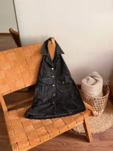 Vestido corto de jean negro con cuello halter, cierre frontal con botones dorados, bolsillos con solapa en el pecho y corte evasé.