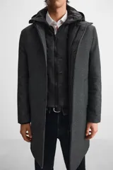 Abrigo gris oscuro de corte recto, con cuello doble y capucha desmontable con cremallera. Tiene manga larga, bolsillos laterales y abertura central en la espalda. El cierre frontal es con cremallera oculta por solapa con botones a presión.