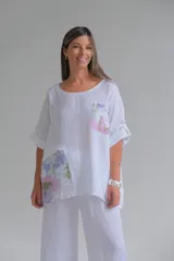 Blusa blanca de lino con cuello redondo y mangas 3/4 con botón. Tiene un bolsillo en el pecho y apliques estampados con motivos florales.