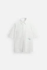 Camisa blanca de algodón con estructura canvas, corte boxy fit, cuello bowling y manga corta. Presenta bordado en azul claro con texto cursivo en el pecho y botones frontales.