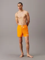 Bañador corto color naranja con cintura elástica ajustable con cordón blanco, bolsillos laterales y logo de Calvin Klein estampado en blanco en la pierna izquierda.