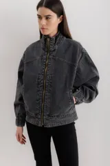 Campera tipo bomber de jean negro con cierre frontal dorado, bolsillos laterales y cuello alto. Tiene mangas largas con puños elastizados y detalles de costuras a contratono en cuello y pretina.