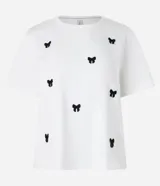 Remera baby look blanca de algodón con cuello redondo y mangas cortas. Presenta bordados de lacitos negros en el frente.